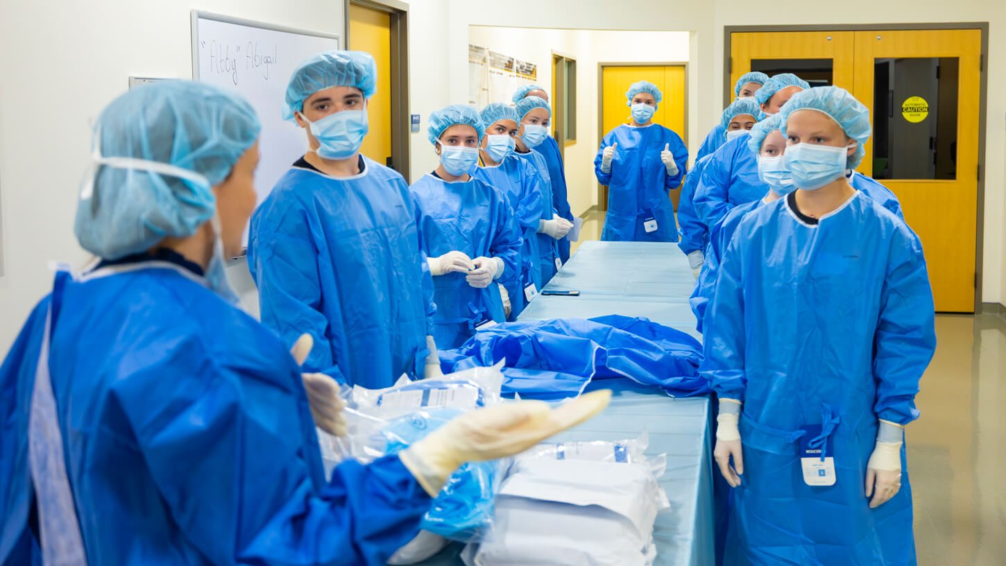A nursing instructor demonstrates a medical procedure on a manikin while a group of students in scrubs watch attentively behind a surgical drape.