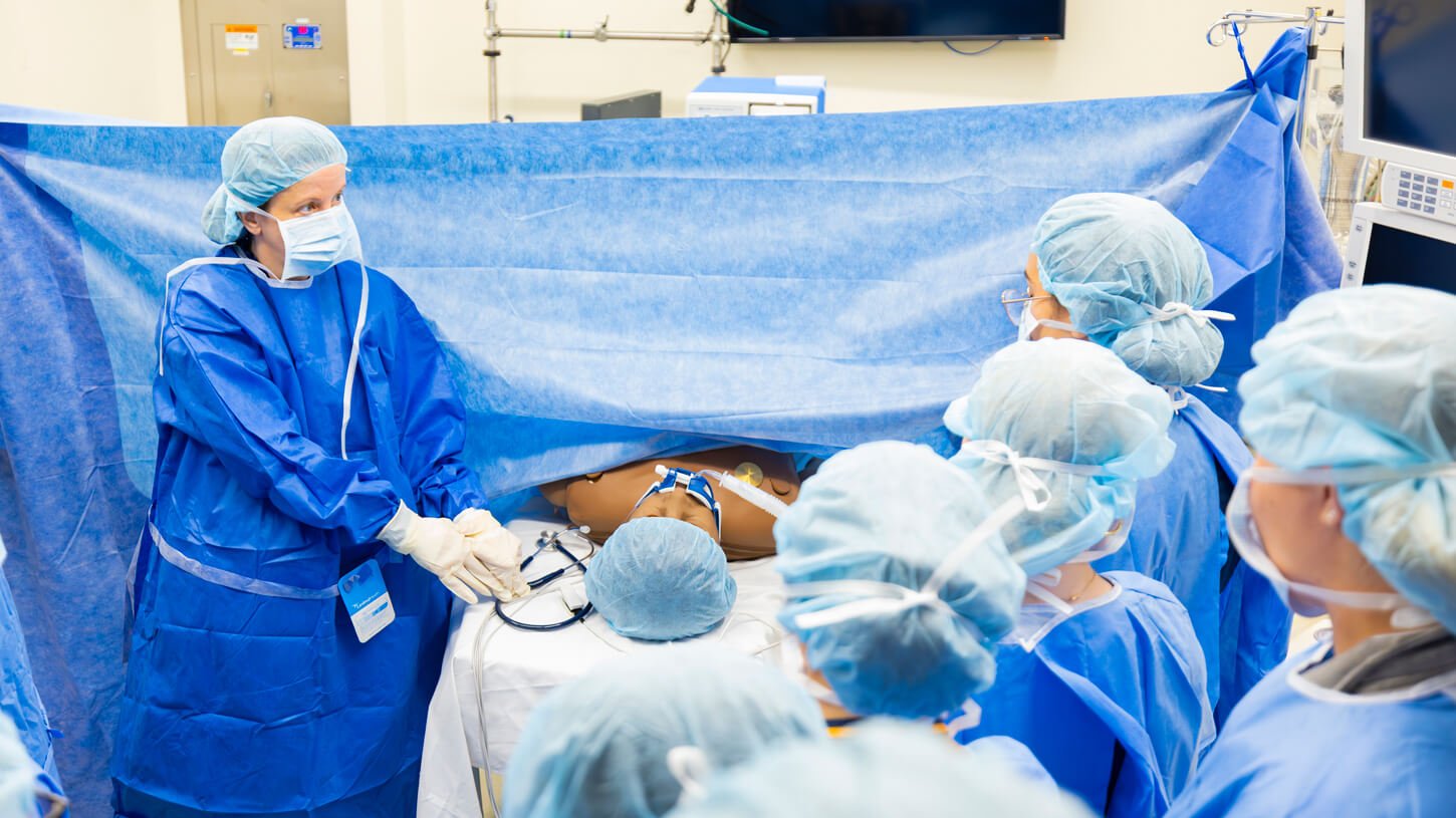 A nursing instructor demonstrates a medical procedure on a manikin while a group of students in scrubs watch attentively behind a surgical drape.