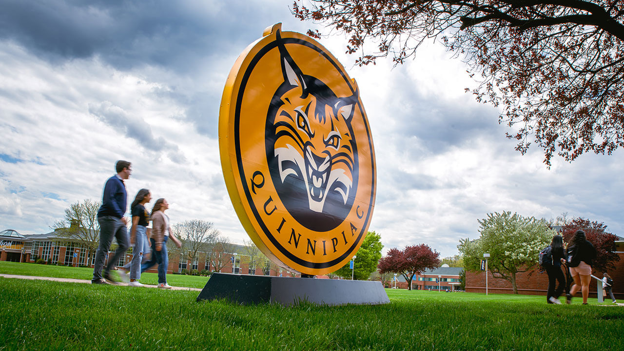 Exterior shot of the Quinnipiac sign while students walk past the Quad