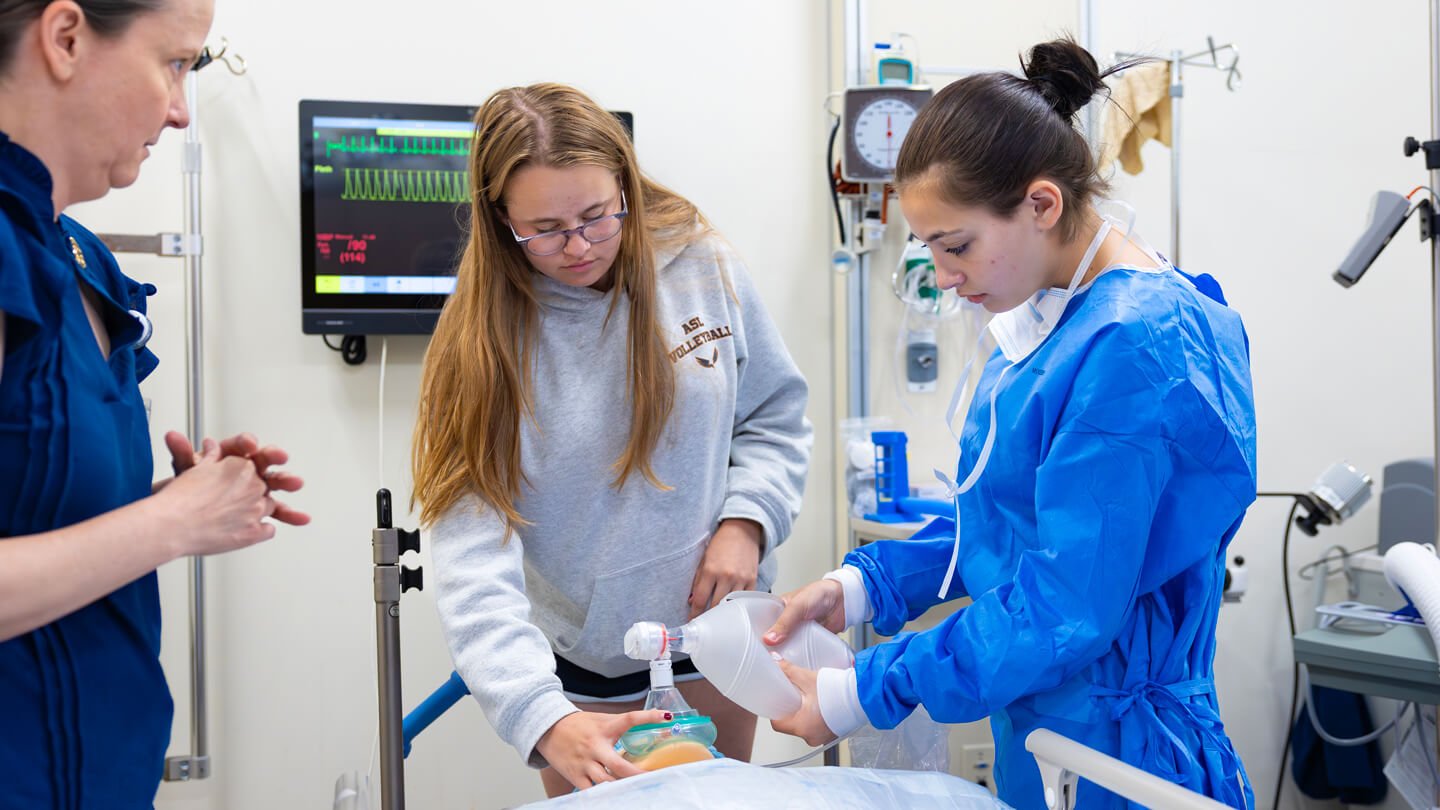 Two students work together to ventilate a mannequin using a bag valve mask as an instructor observes nearby.