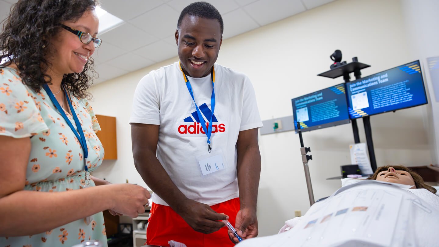 A student smiles while learning to administer an injection to a medical mannequin, guided by an instructor during a simulation activity.