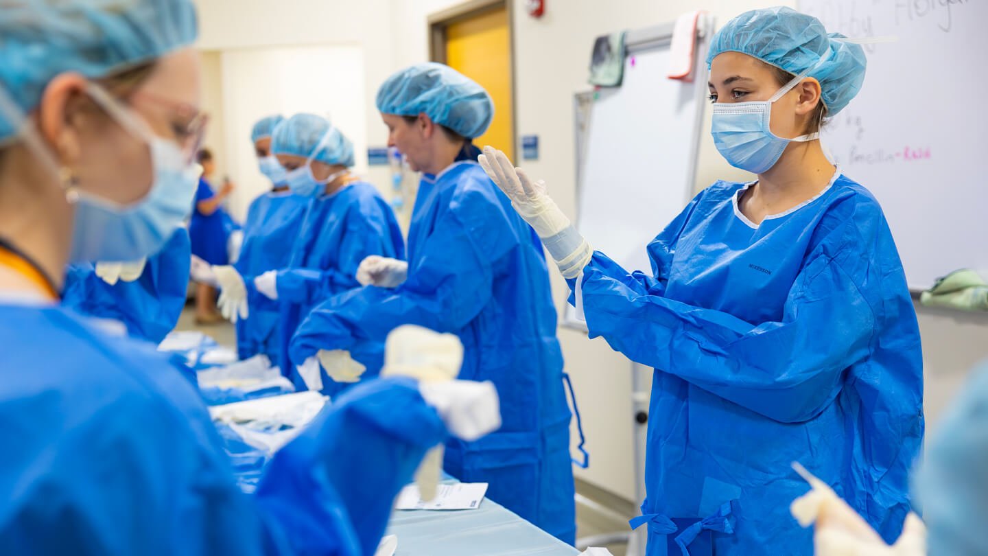 High school students in blue surgical gowns and masks practice sterile glove techniques during a nursing summer camp session at Quinnipiac University.