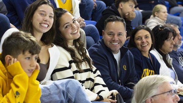 Smiling students and their families in Quinnipiac gear smile in the ice hockey stadium stands