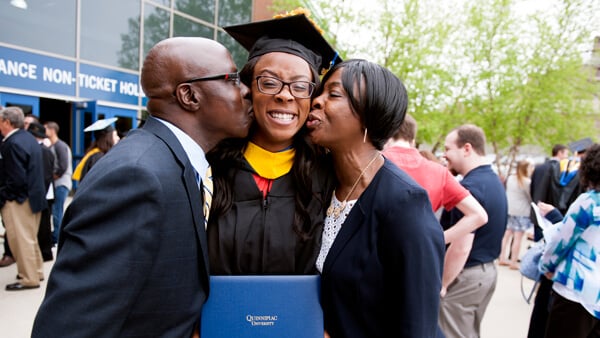 A Quinnipiac graduate holds her diploma and smiles while her parents kiss her on the cheeks