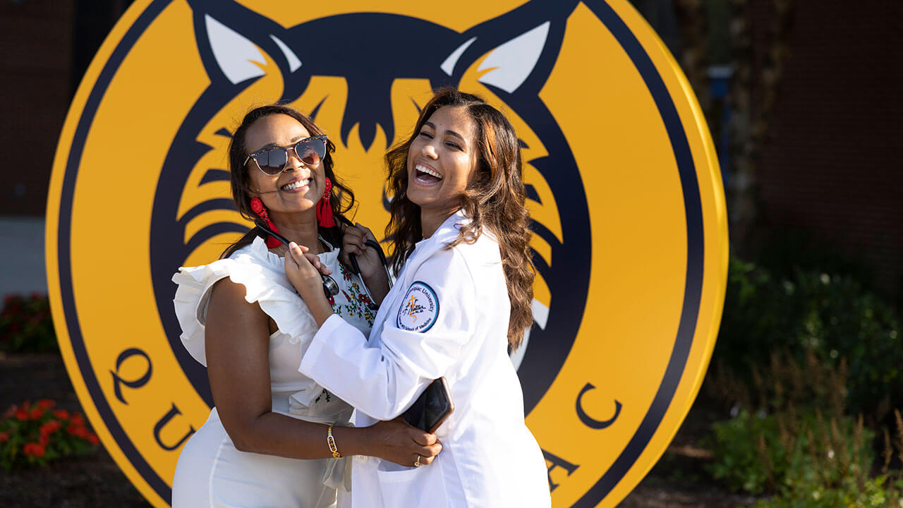 Two graduates take a photo in front of a Quinnipiac sign
