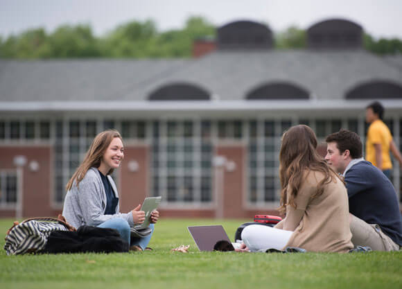 Students sit with laptops on the quad grass in front of the library