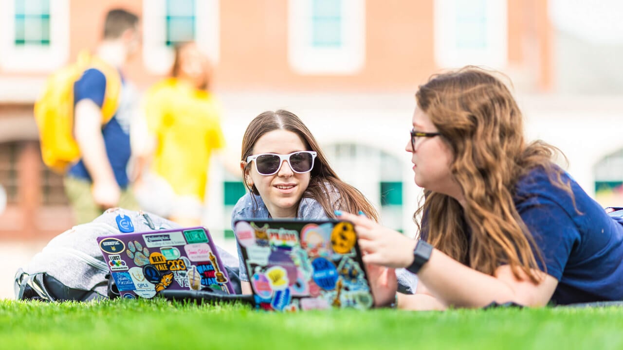 Two students lay on the grass on campus with open laptops studying