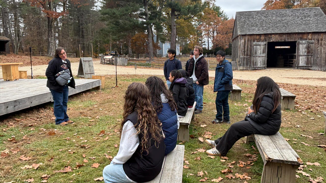 A faculty member speaking to a group of students sitting on benches outdoors.