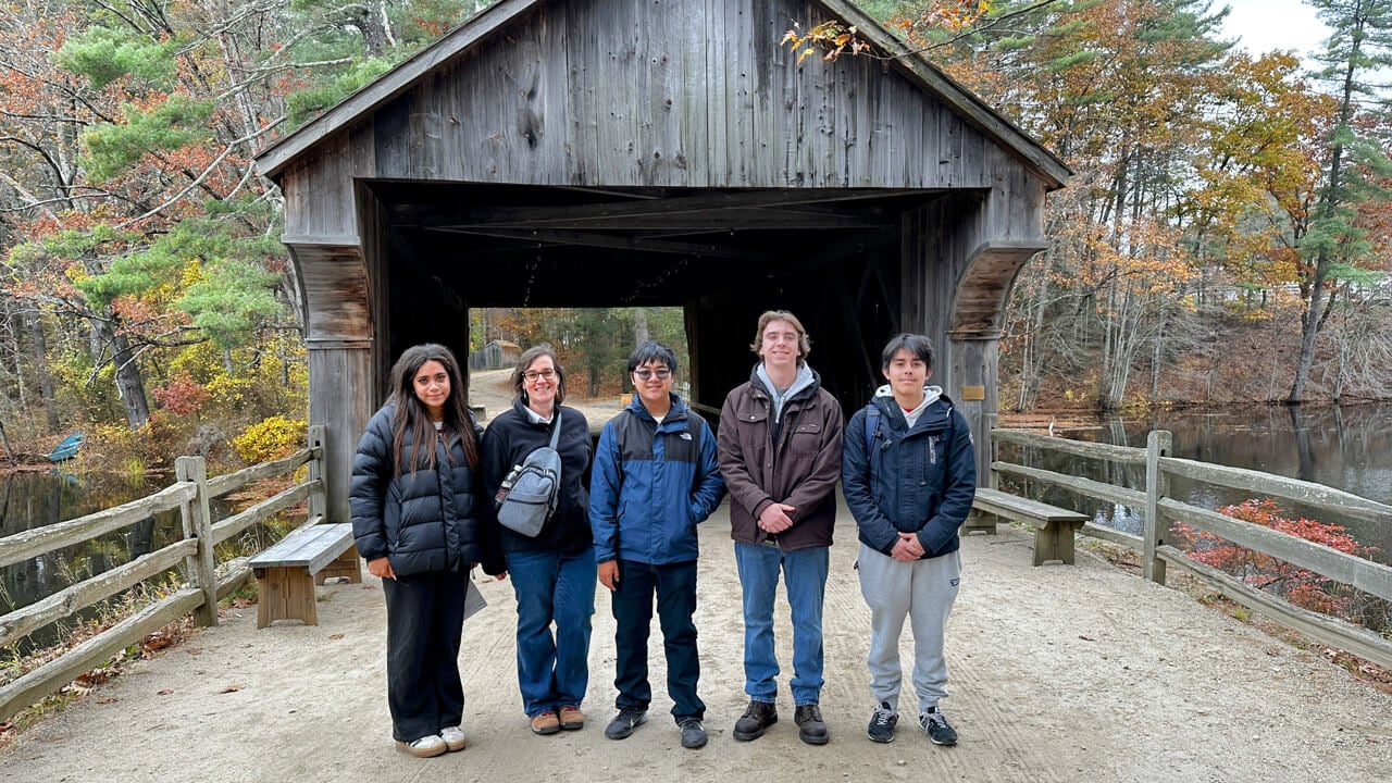 A group of five participants standing in front of a covered bridge surrounded by fall foliage.