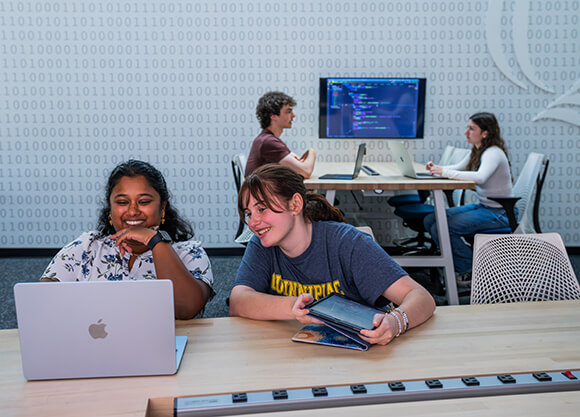 Two students on laptops in a CCE classroom. A blurry background shows two more students, one of them pointing at a screen on the wall.