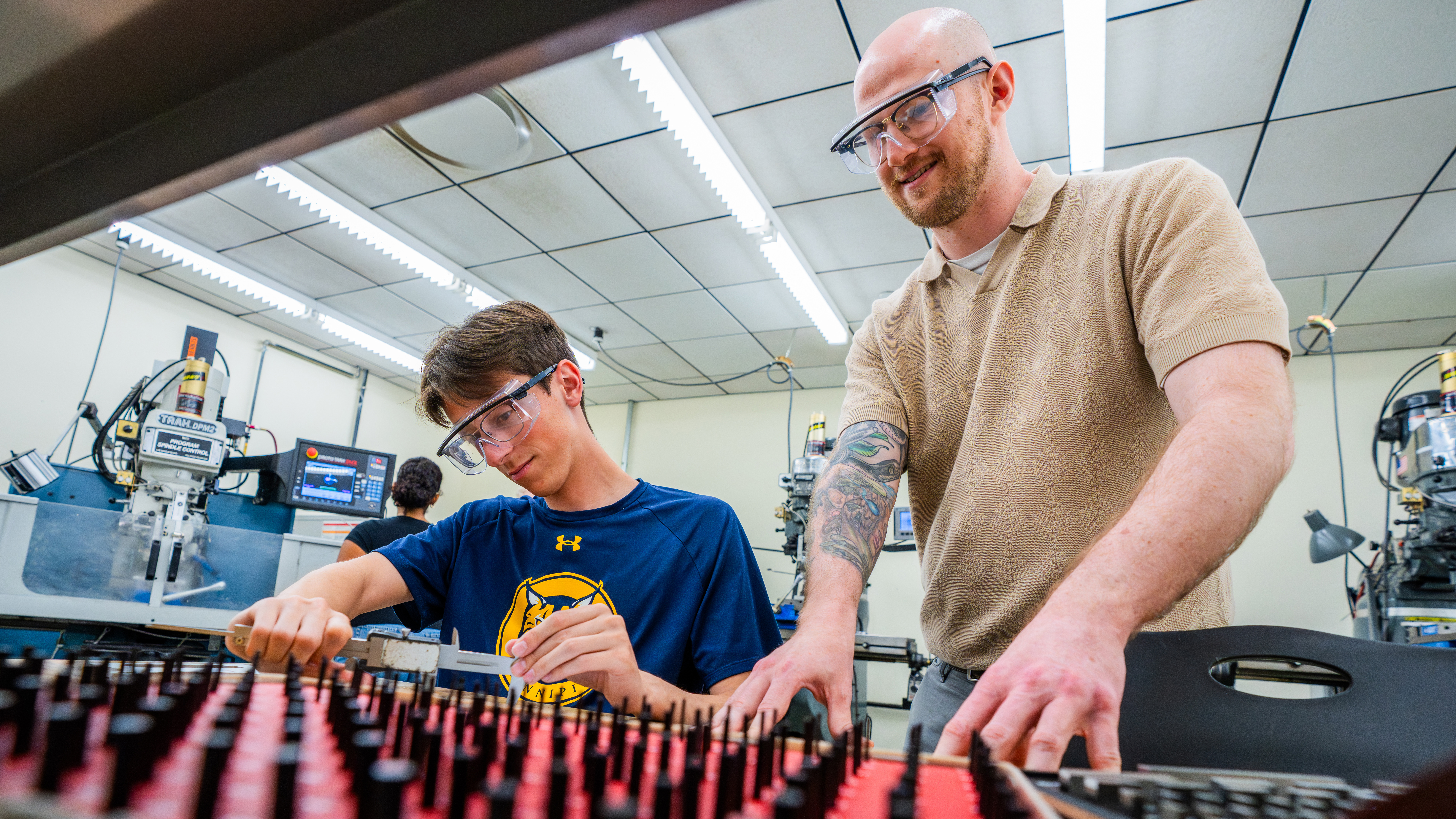Two male students in safety goggles using red and black switchboard-like equipment.