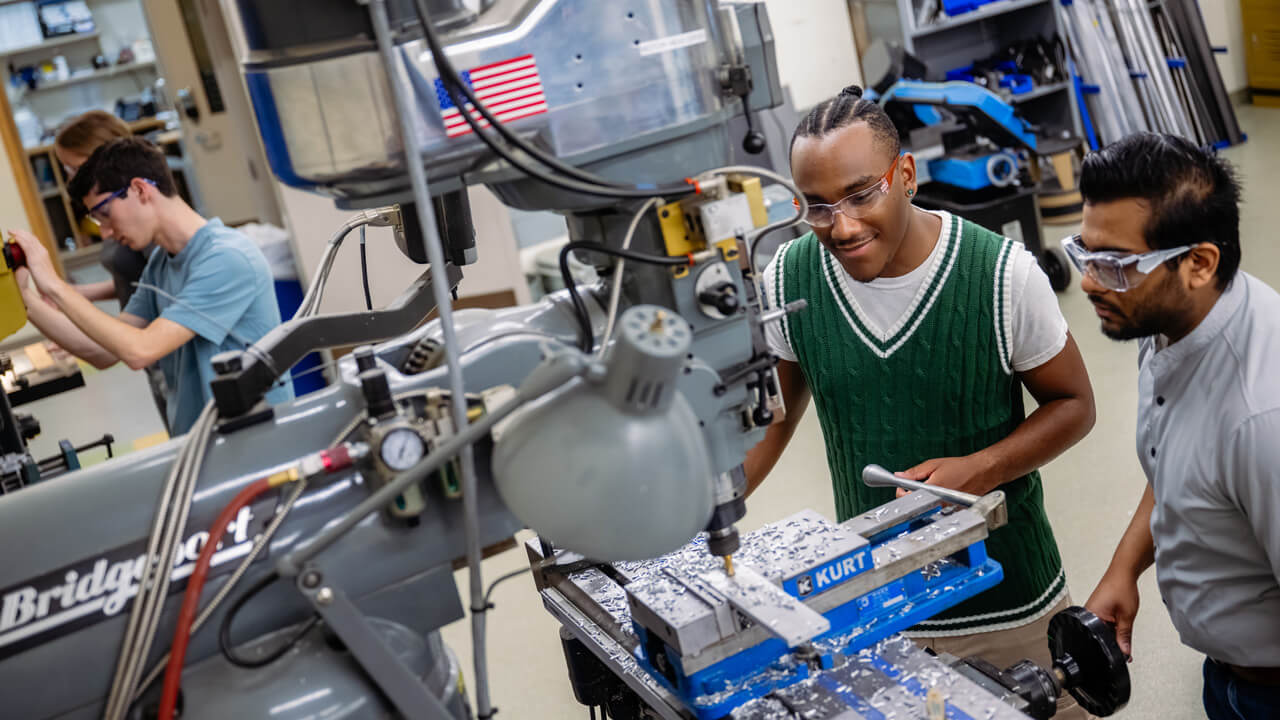 Students and faculty in safety goggles looking at machine shop equipment labeled "KURT."