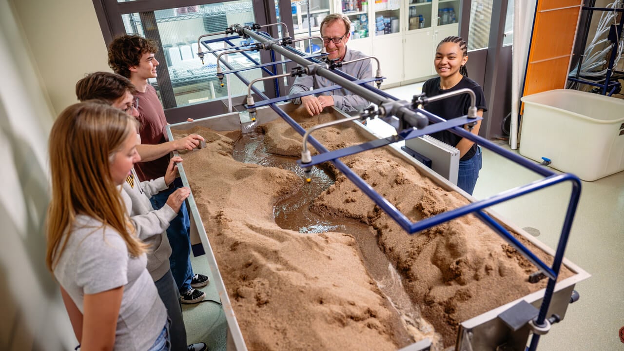 A shot from above of four students and a faculty member looking into a basin in the Fluids Lab filled with soil and water.