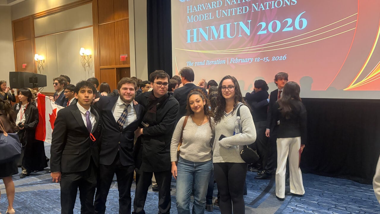 Five students in a busy conference room posing in front of a "Harvard National Model United Nations 2026" slideshow.