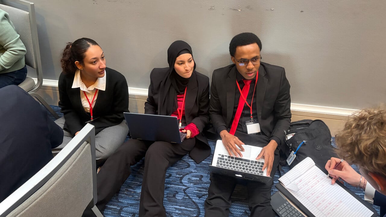 Three students sitting on the floor of a hallway with laptops. Someone else is shown from behind writing on a pad of paper.
