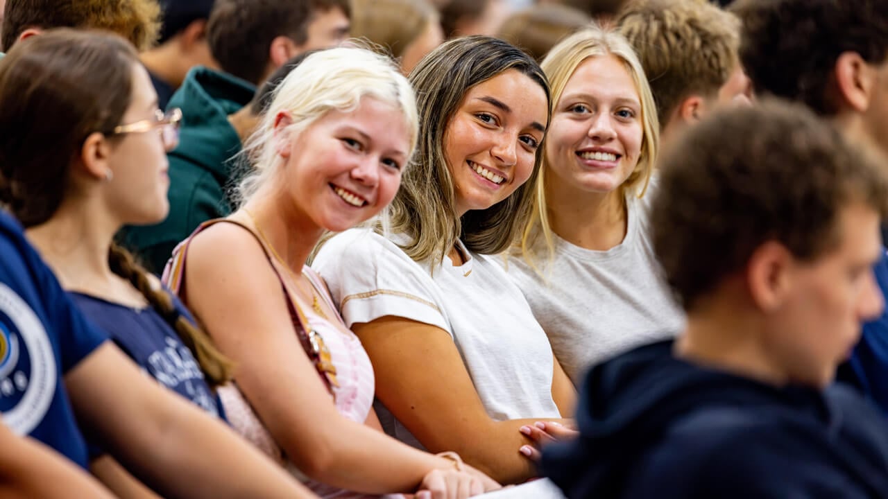 Three students smile as they sit together among their new student peers in the recreation center