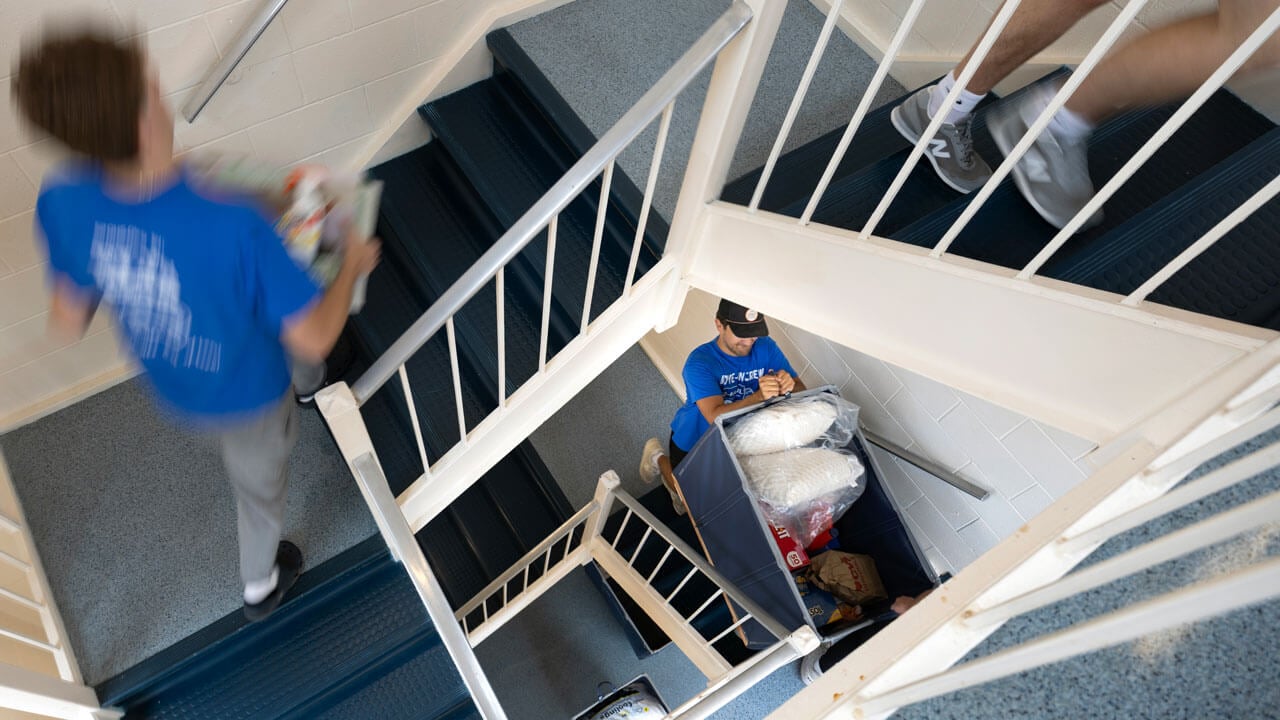Students cary items up the stairs in a dorm building.
