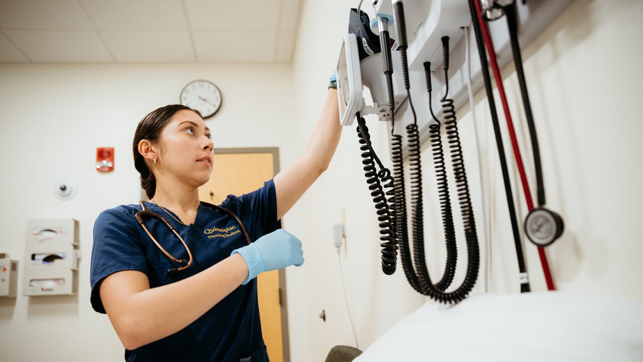 A Quinnipiac nursing student in scrubs adjusts medical equipment on the wall of a simulation lab, preparing for hands-on training.