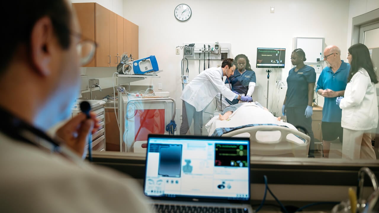 Nursing students perform an exam on a manikin patient in a simulation suite with faculty giving direction from an observation room.