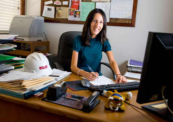 Holly LaPrade pauses to smile while working at her desk that includes a small stuffed QU Bobcat