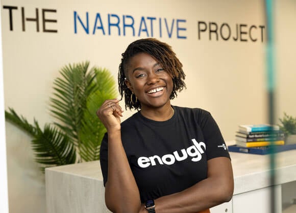 Mercy Quaye wearing a  t-shirt that says "enough" stands in front of The Narrative Project office sign
