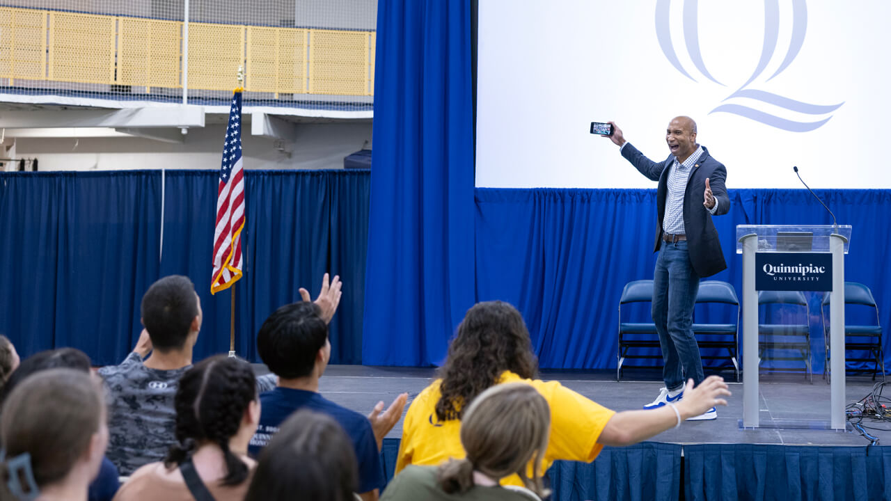 Austin Ashe holds up his phone on stage and encourages students to do the Quinnipiac Bobcat cheer