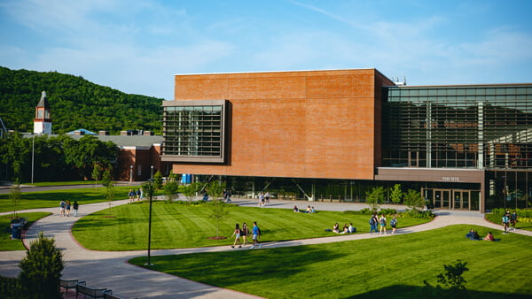 Students walk on paths and sit on grass in front of The SITE with the library clocktower in the background