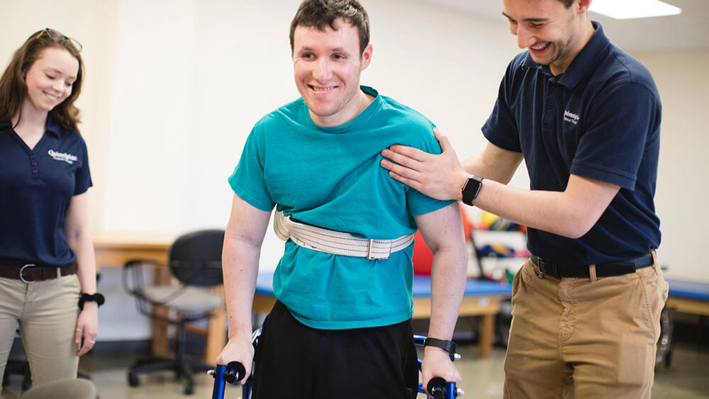 Two health sciences students stand on either side of a patient using a walker and help him stand up