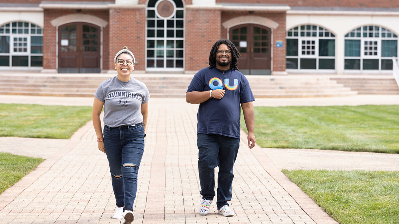 Two students in Quinnipiac students walk in front of the Arnold Bernhard Library