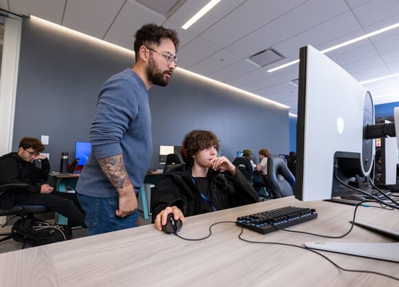 A student clicking a mouse looking at a monitor while a faculty member stands next to him looking on.