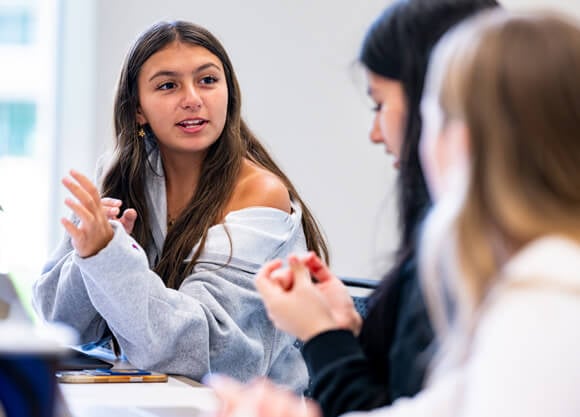 A female student sitting at a desk talking to other students in the foreground who are blurry.