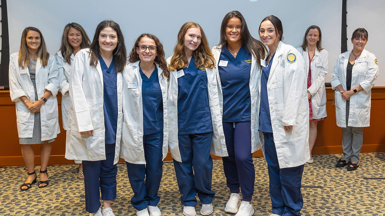 five nursing students pose smiling in their new white coats in front of their advisors