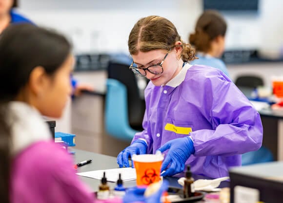 A female student in a purple smock and rubber gloves examines a sample.