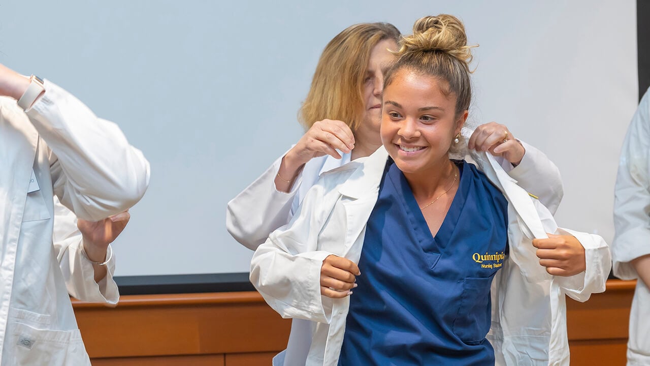 smiling nursing student puts her white coat on with the assistance of a woman behind her