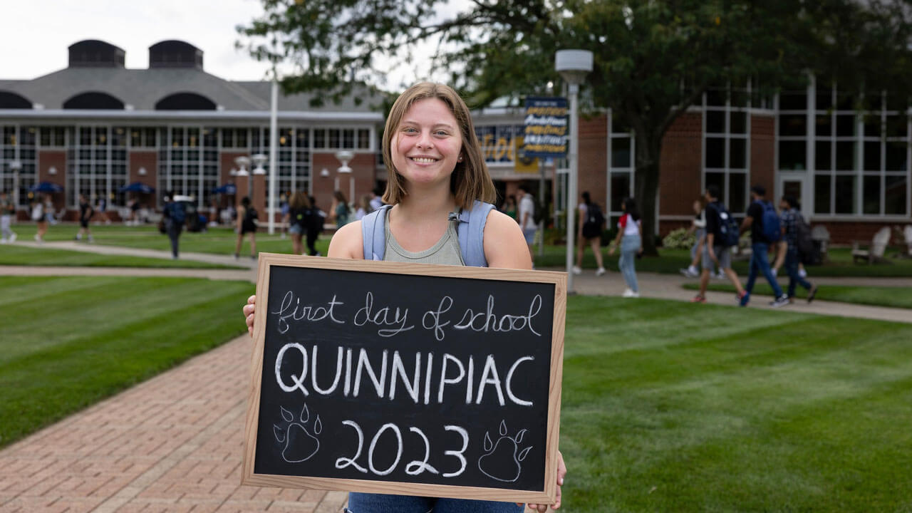 Student smiles while holding first day of classes written on chalkboard
