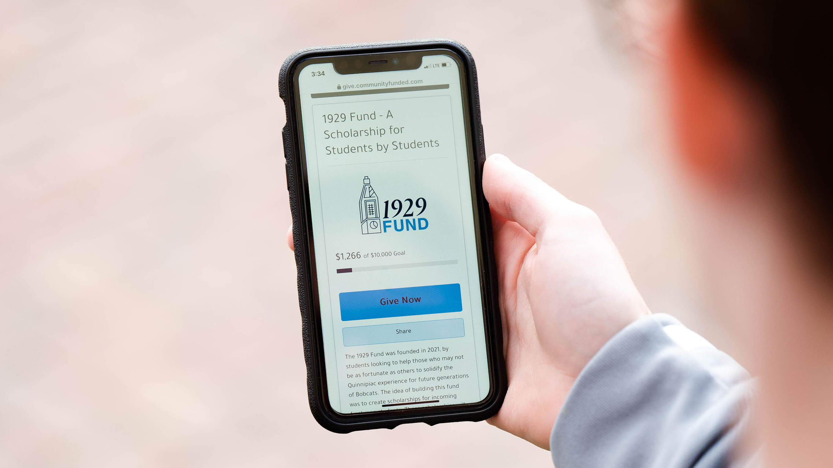 Someone's hand holding an iPhone with a blurred out background. On the screen is the 1929 Fund donation webpage.