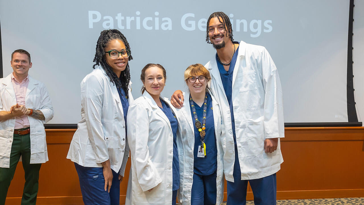 four nursing students in white coats pose smiling for a photo