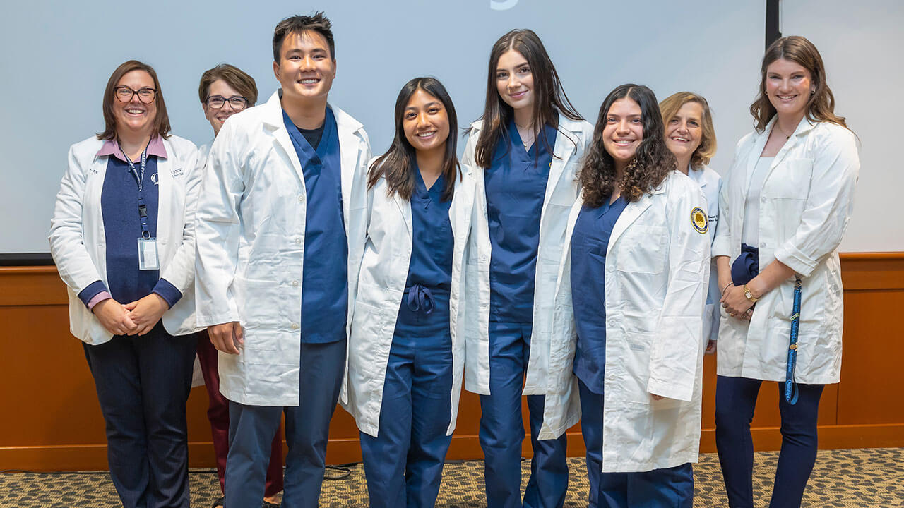 four nursing students pose in their white coats in front of their advisors