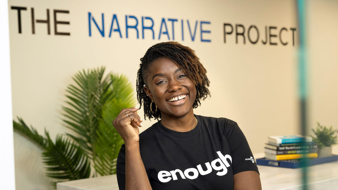 Mercy Quaye wearing a t-shirt that says "enough" stands in front of The Narrative Project office sign