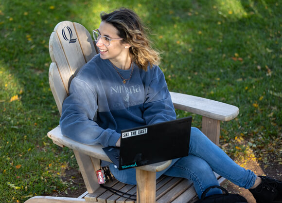 A student smiles and looks over her shoulder while she sits in an Adirondack chair on the quad with her laptop