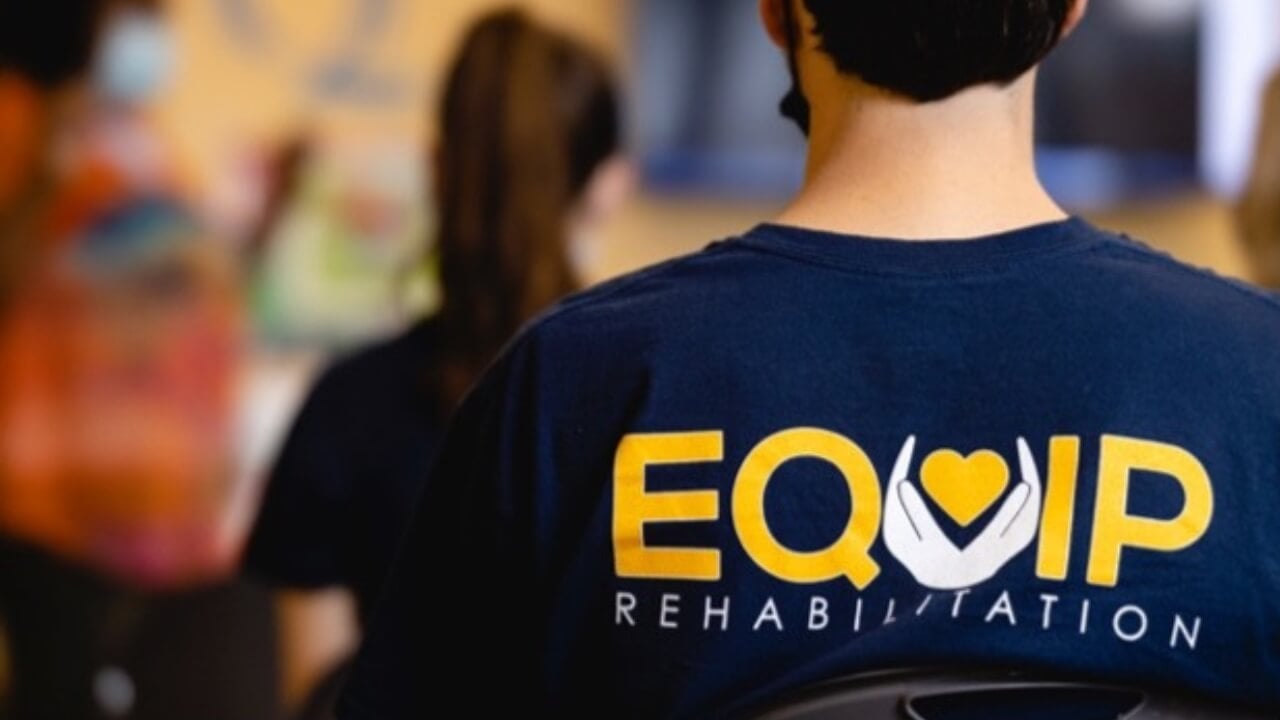 A photo of the back of a male that is wearing a navy blue t-shirt that says Equip Rehabilitation