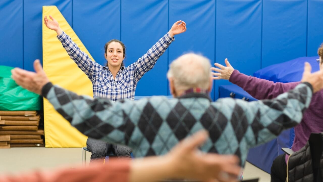 A photo of woman with her arms up leading an exercise session for elderly folks.
