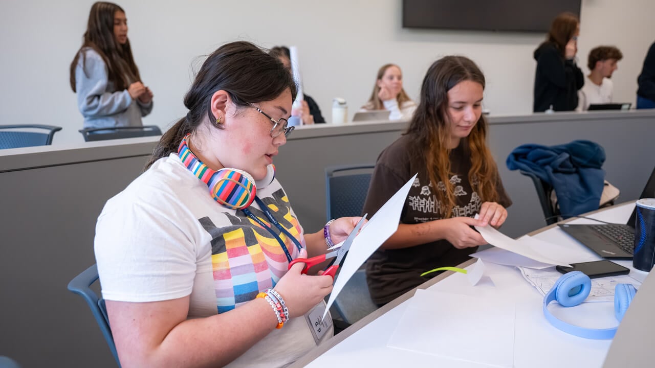 Two students at a desk cutting pieces of paper. One has a large pair of rainbow headphones around her neck.