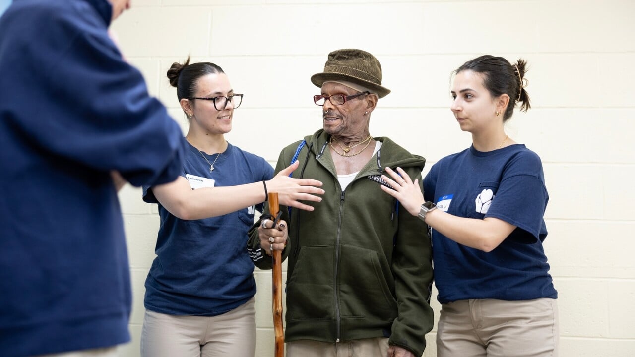 A photo of two female students assisting an elderly man with a cane.