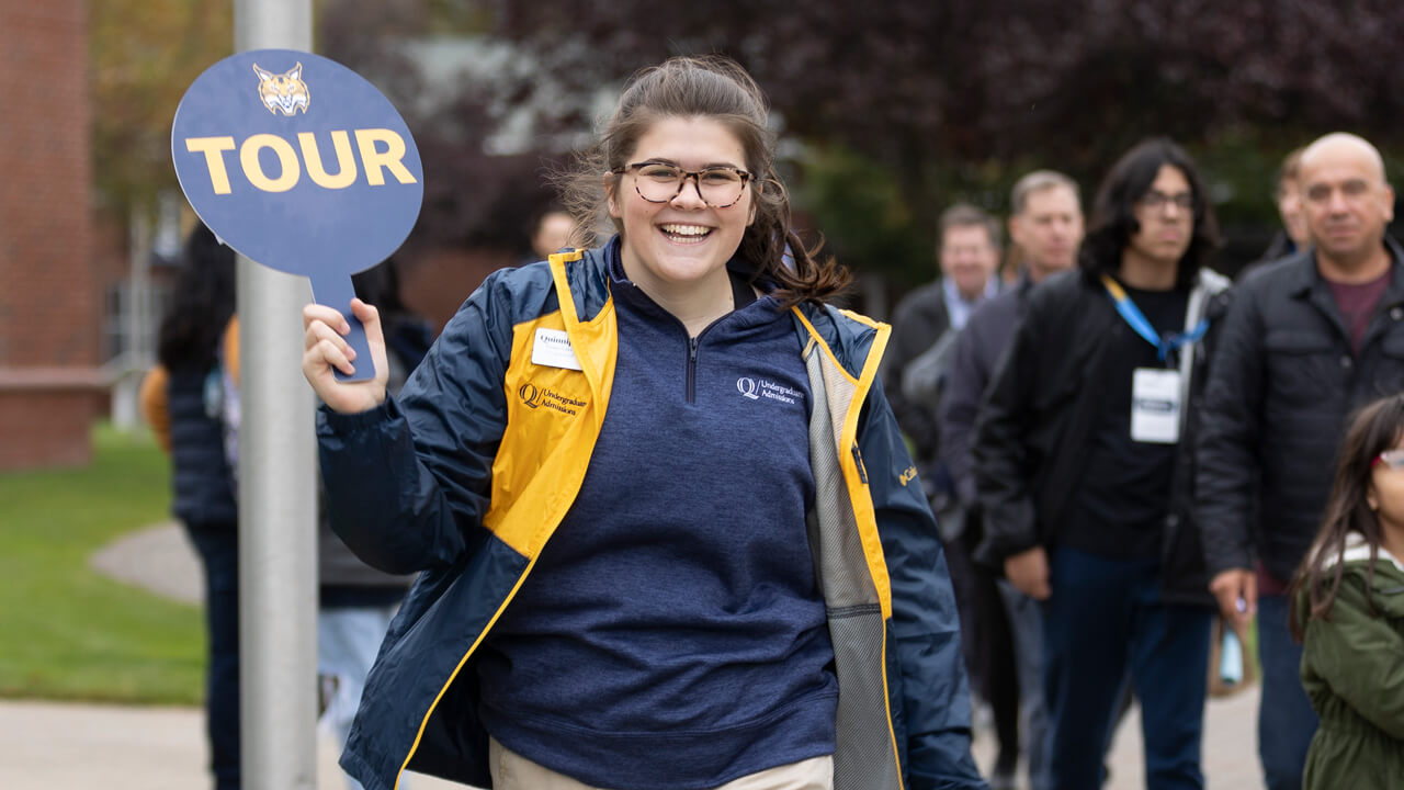 An admissions student guide in Quinnipiac colors smiles as she holds up a tour sign