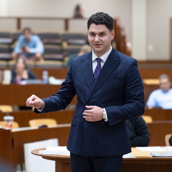 A law student in a suit gesturing as he argues in a courtroom.
