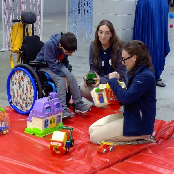 A participant using a wheelchair and two staff members sit together on a red mat during a play-based therapy activity. Toy houses, vehicles, and colorful play equipment are arranged on the mat in an indoor therapy space.