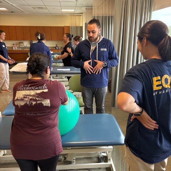 Rehabilitation instructor demonstrates a therapy exercise using a stability ball while participants practice in a clinical training setting.