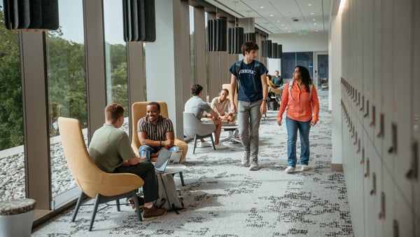 Students sit in chairs by a wall of windows as other students walk by