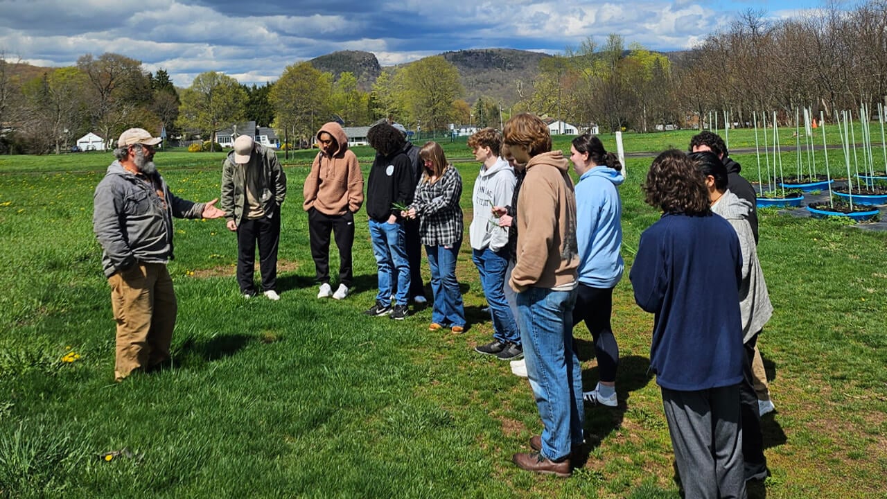 A male faculty member talking to a group of students standing in a line in a green field.